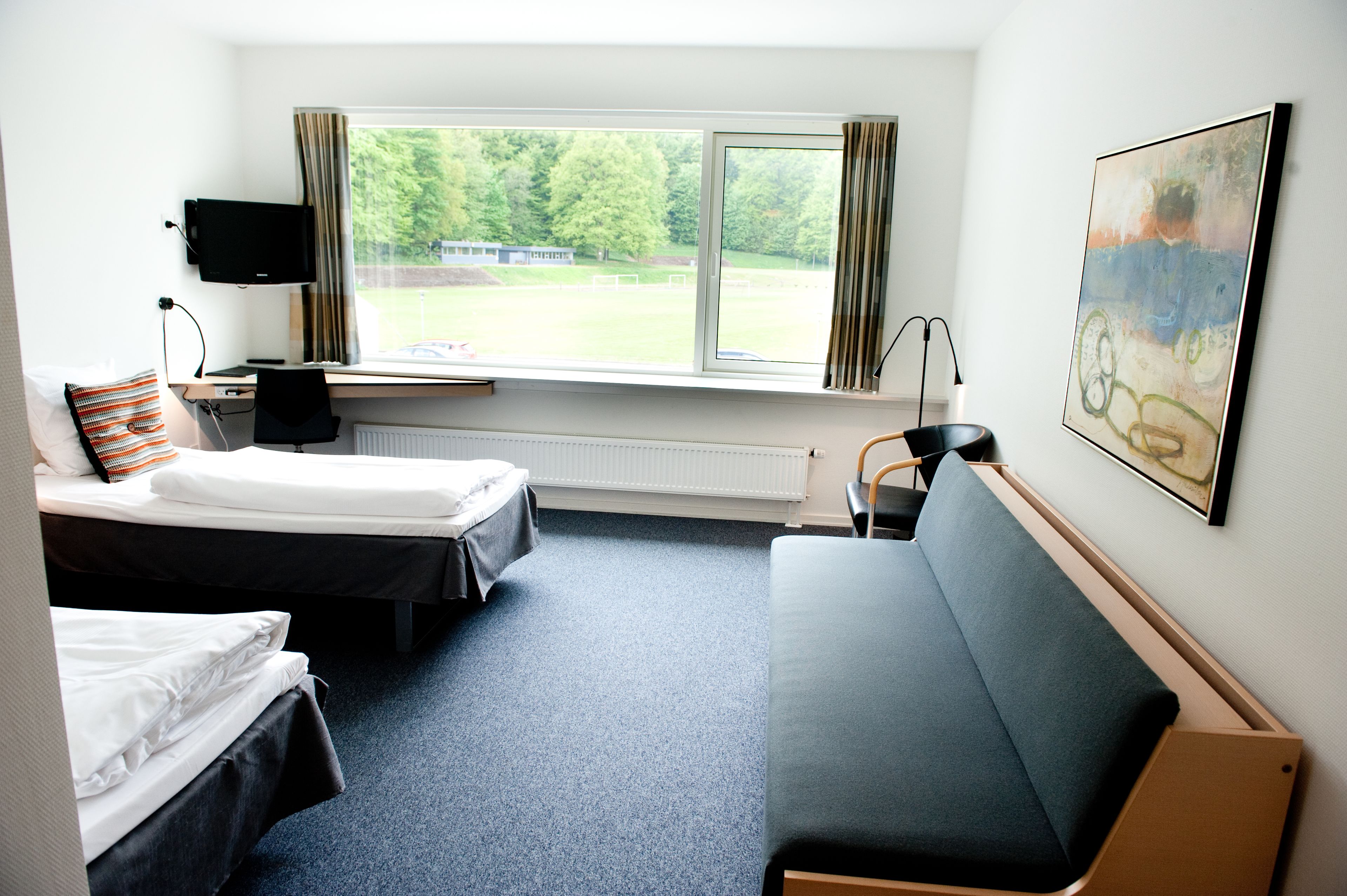 A hotel room with two beds, a desk with a computer, a chair, a couch, a window with a view of a green landscape, and a painting on the wall.