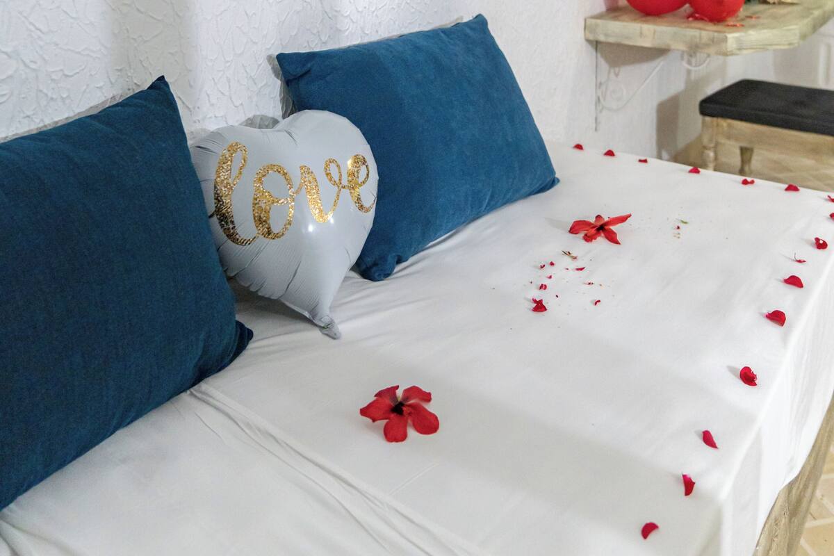 A bed with blue pillows, a white pillow with 'Love' written on it, and red rose petals scattered on the bed.