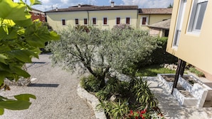 Un cortile con un vialetto sterrato, un piccolo giardino con diverse piante, e un edificio con molte finestre e un camino.