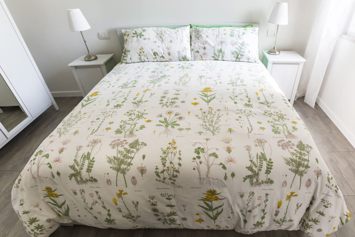 En pænt redt seng med blomstret sengetøj, to sengeborde med lamper, og en hvid væg.
