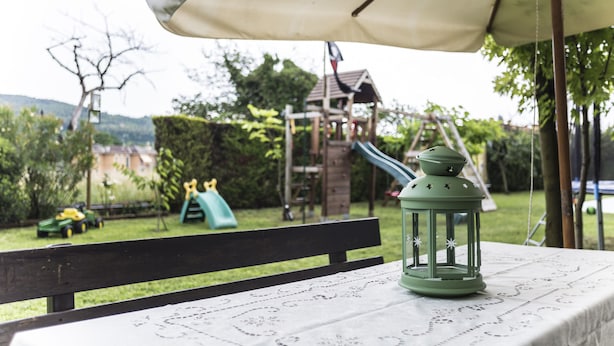 Un tavolo con una lanterna verde e una tovaglia bianca, sistemato sotto un grande ombrello in un giardino con un'area giochi sullo sfondo.