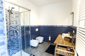 A modern bathroom with a glass shower enclosure, two toilets, a wooden vanity, and a black bath mat.
