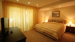 A hotel room with a large bed, a desk with a computer and a remote control, a nightstand with a lamp, and a painting on the wall. - Vox Maris Grand Resort - Adults only (Costinesti)