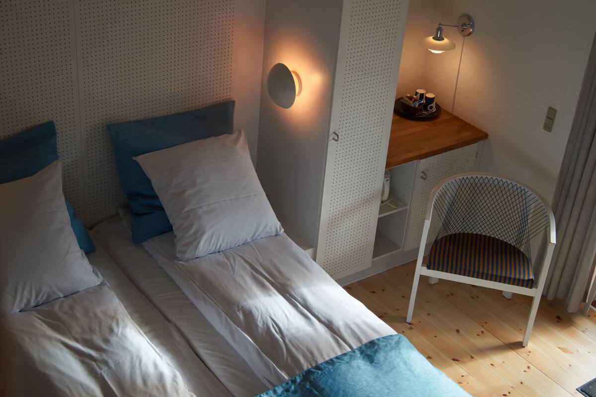 A bedroom with two beds, a chair, a desk, and wall-mounted lighting.