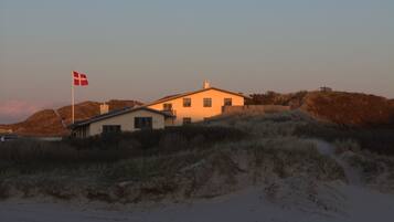A coastal house with a flag, surrounded by dunes and a beach.