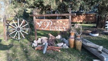 Une enseigne rustique en bois, ornée du mot « PUALY » et d’éléments décoratifs, entourée d’un aménagement paysager naturel et d’une grande roue en bois.