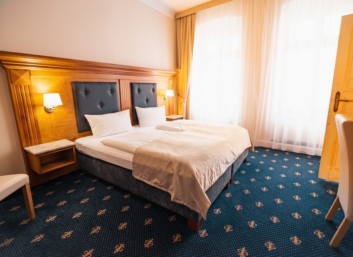 A hotel room with a large bed, two bedside tables, a nightstand, a wooden headboard, and a window with curtains.