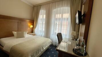 Ein Hotelzimmer mit einem großen Bett, einem Schreibtisch mit Stuhl, einem an der Wand befestigten Fernseher und einem Fenster mit Vorhängen.