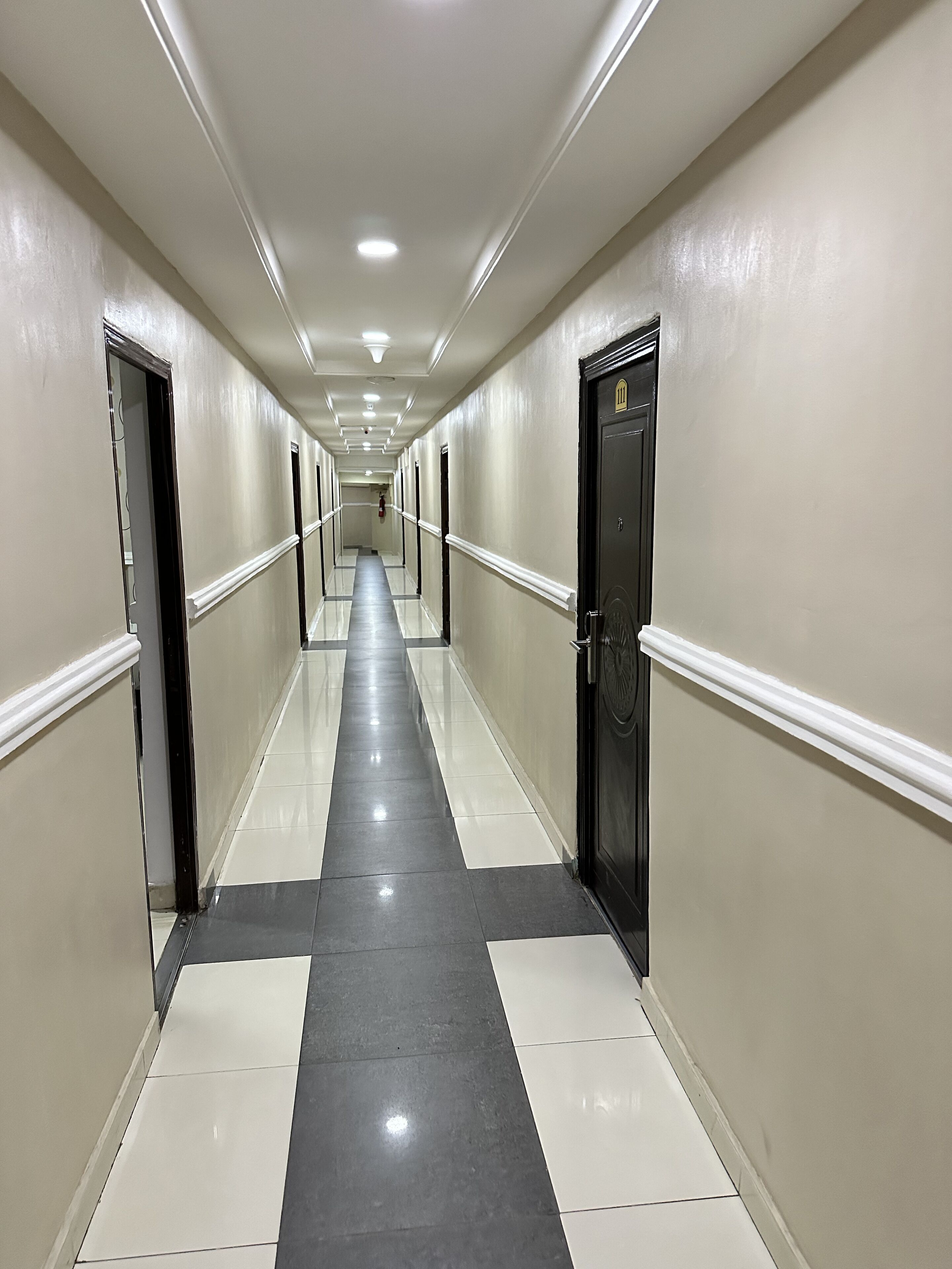 A long, narrow hallway with a reflective black and white tiled floor, white walls, and doors on both sides.