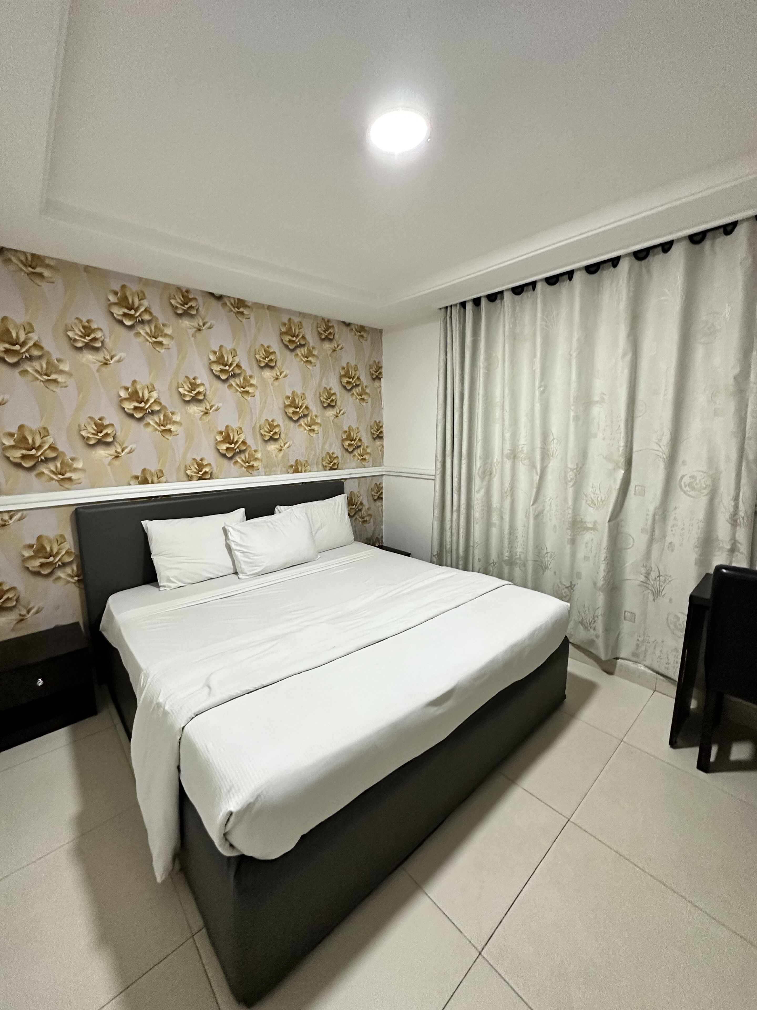 A neatly made bed with white linens in a room with a floral wallpaper, a ceiling light, and a curtain with a subtle pattern.