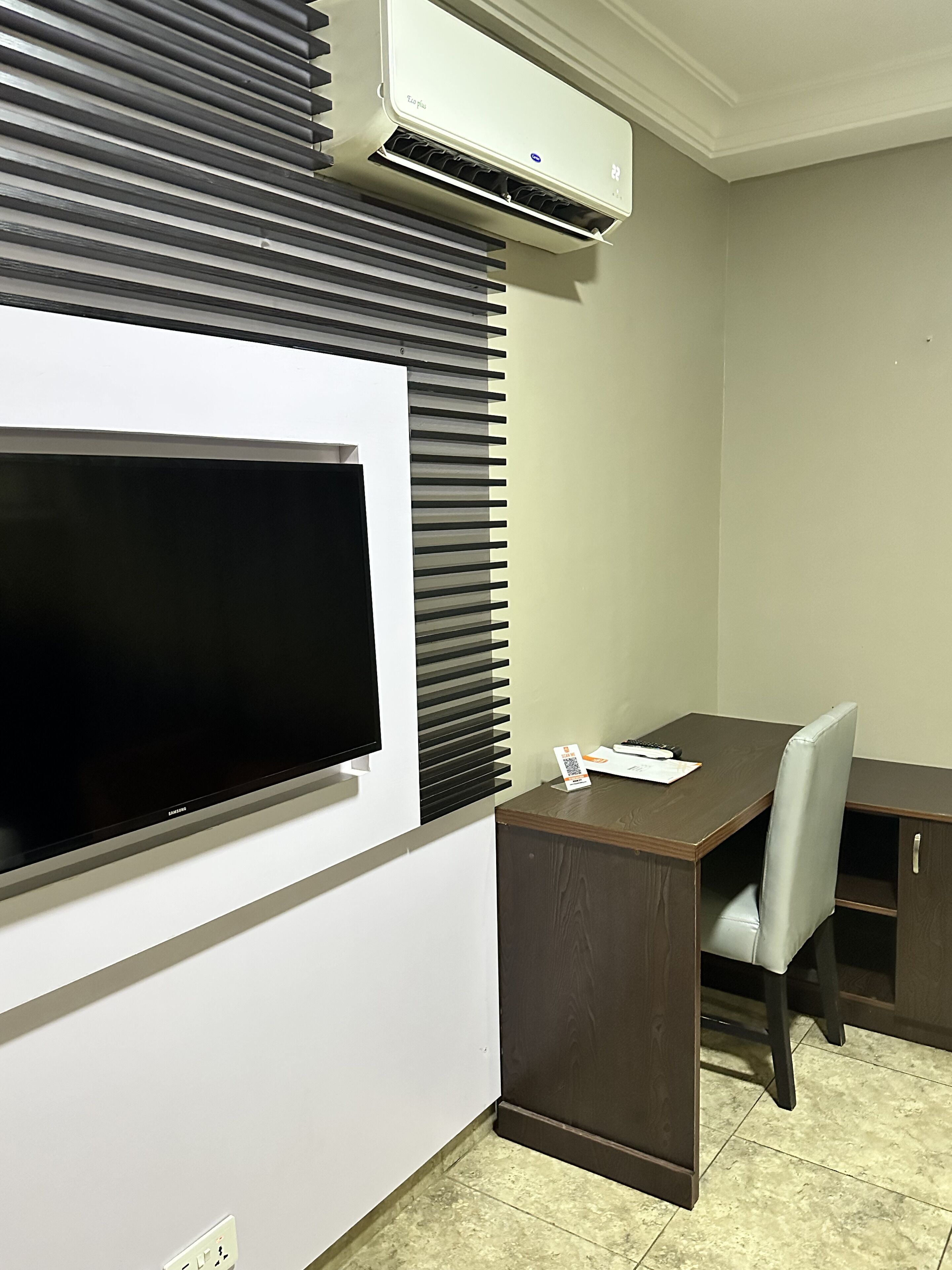 A room with a flat-screen TV mounted on the wall, a wooden desk, a chair, and an air conditioning unit above the blinds.