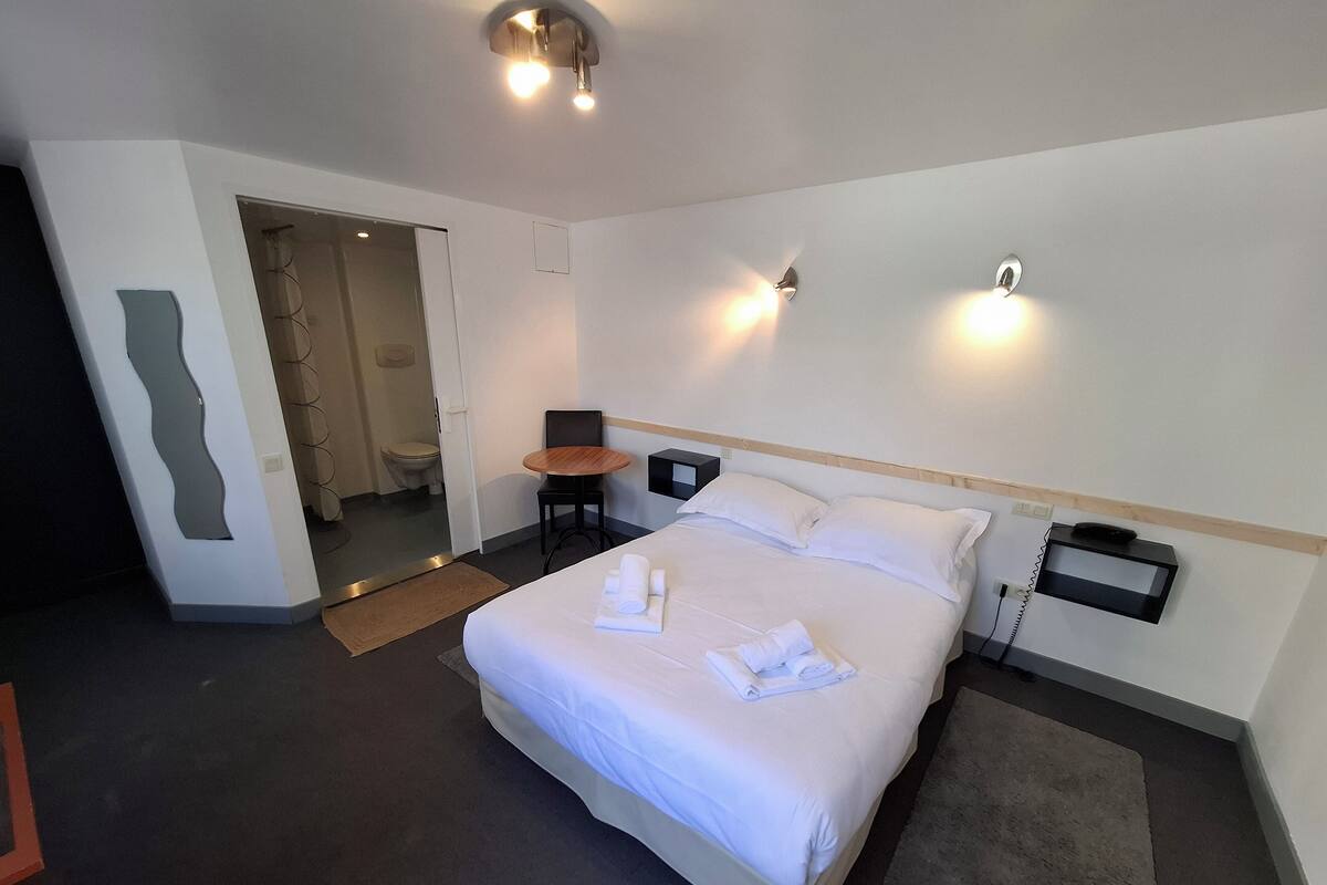 Une chambre d’hôtel avec un grand lit, une petite table, une chaise, une table de chevet avec un téléphone et un miroir au mur.