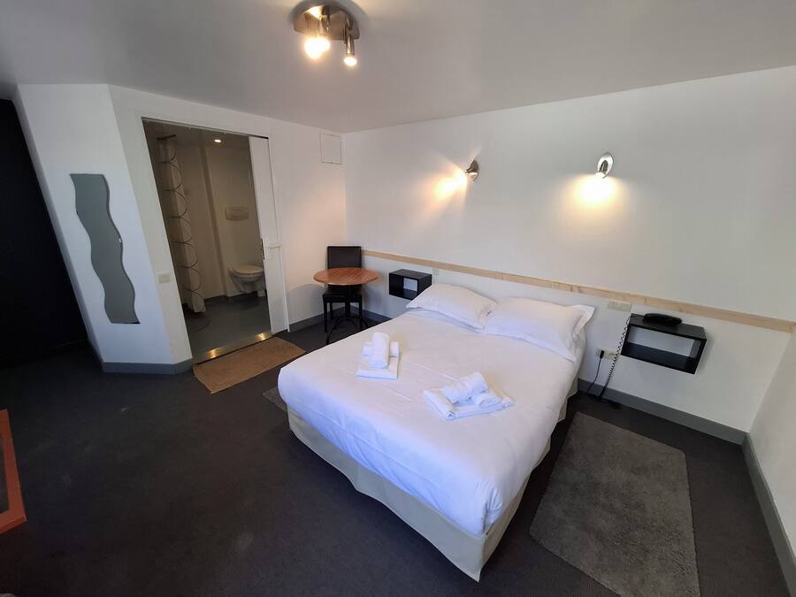 A hotel room with a large bed, a small table, a chair, a bedside table with a phone, and a mirror on the wall.