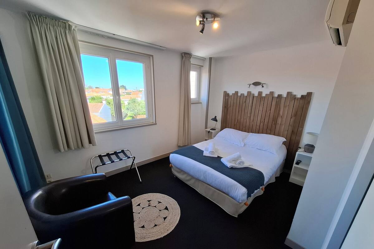 Une chambre d’hôtel avec un lit, une fenêtre avec des rideaux, une table de chevet avec une lampe et une petite chaise.