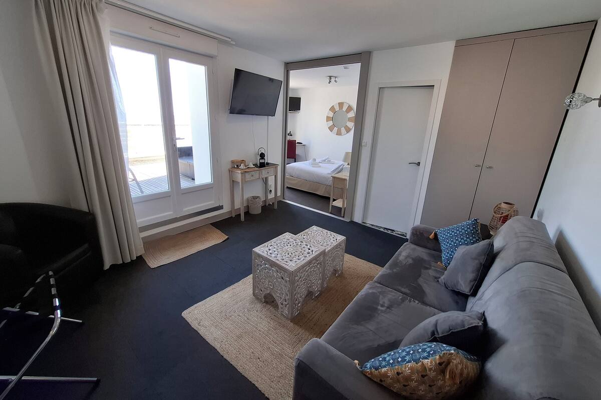 Un salon moderne avec un canapé gris, une table basse et un téléviseur fixé au mur. Une porte donne accès à une chambre à coucher qui contient un lit et une petite table.