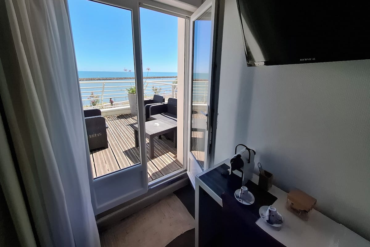 Une pièce dotée d’une grande porte-fenêtre coulissante donnant sur un balcon aménagé avec des sièges et offrant une vue sur la mer.