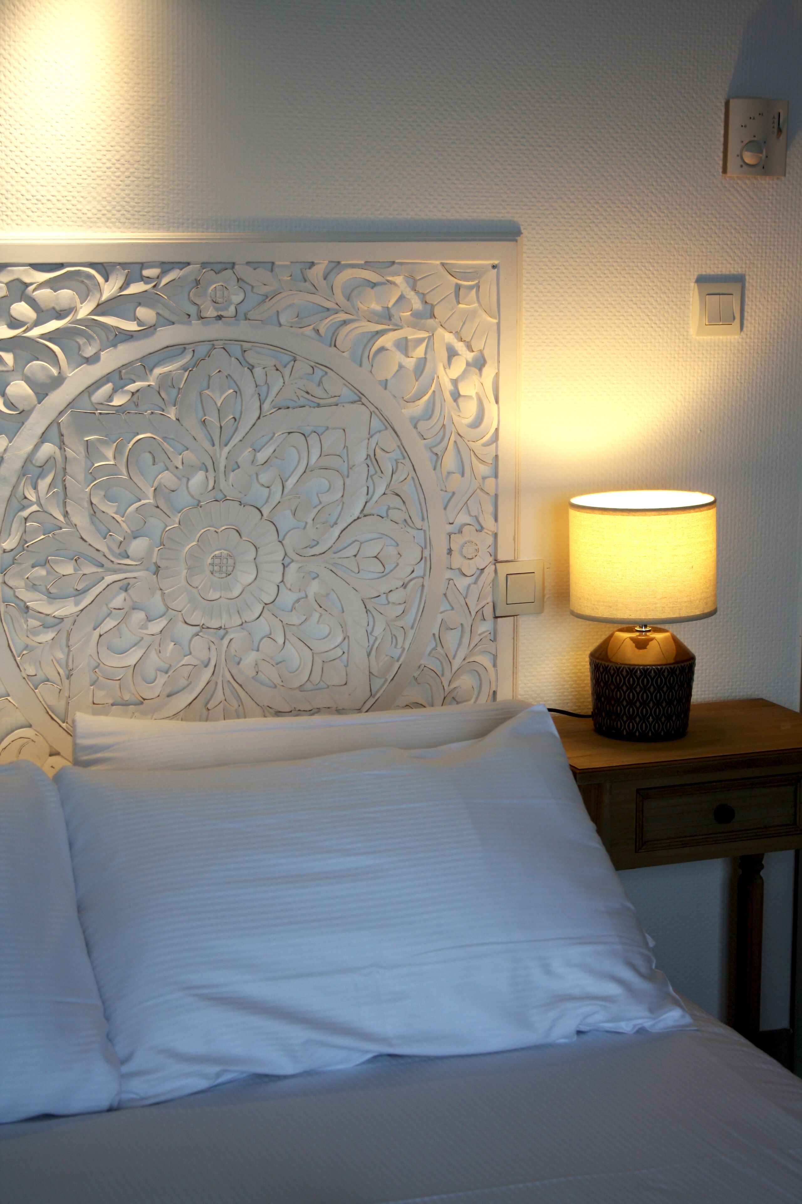 A neatly made bed with white linens, a decorative headboard with floral patterns, a bedside lamp, and a small wooden nightstand.
