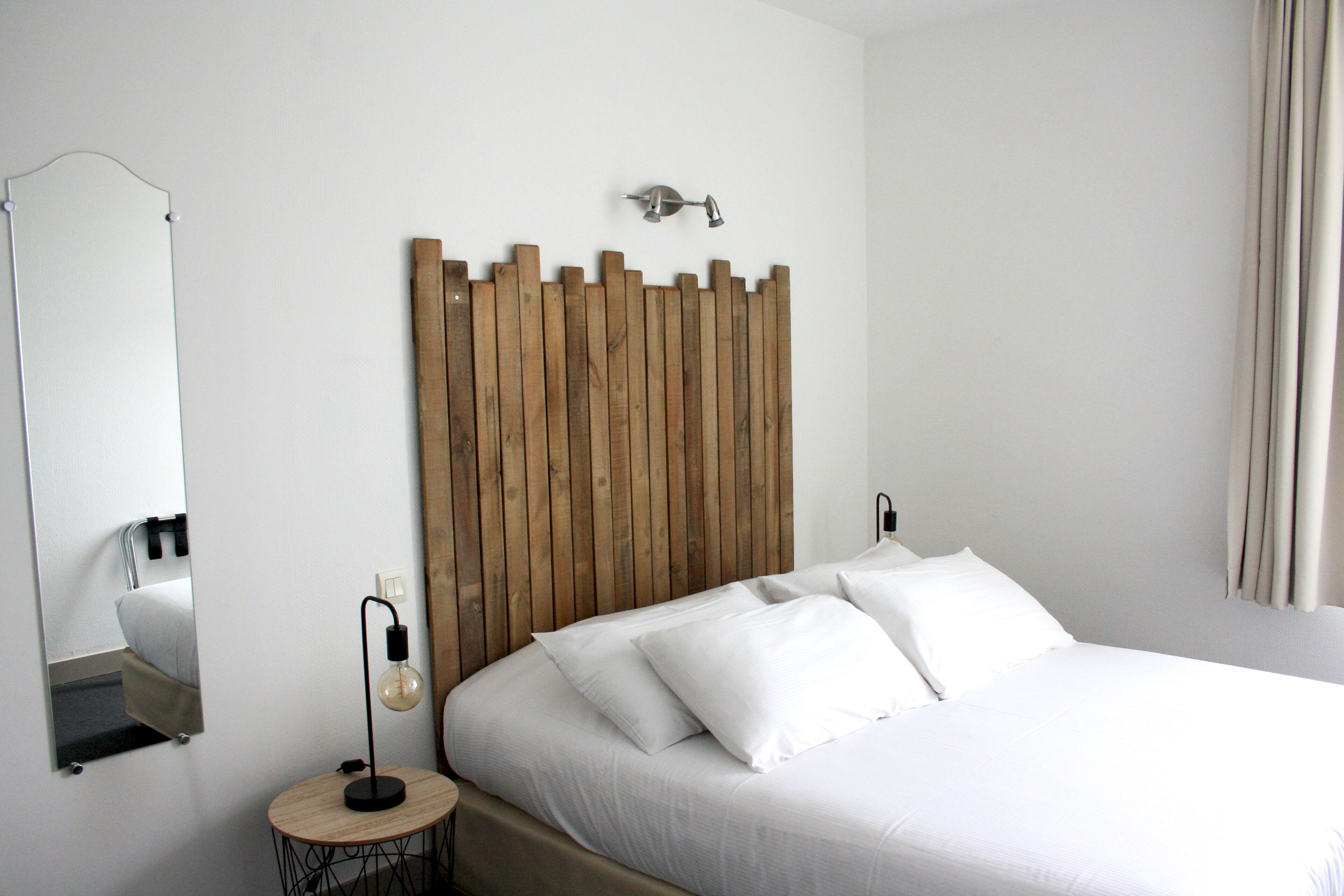 Ein ordentlich bezogenes Bett mit weißen Leinen, ein Holzkopfende, ein runder Nachttisch mit Lampe und ein Spiegel an der Wand.