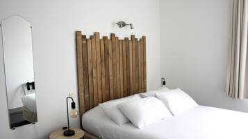Ein ordentlich bezogenes Bett mit weißen Leinen, ein Holzkopfende, ein runder Nachttisch mit Lampe und ein Spiegel an der Wand.