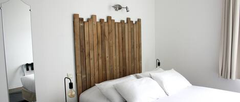 A neatly made bed with white linens, a wooden headboard, a round bedside table with a lamp, and a mirror on the wall.