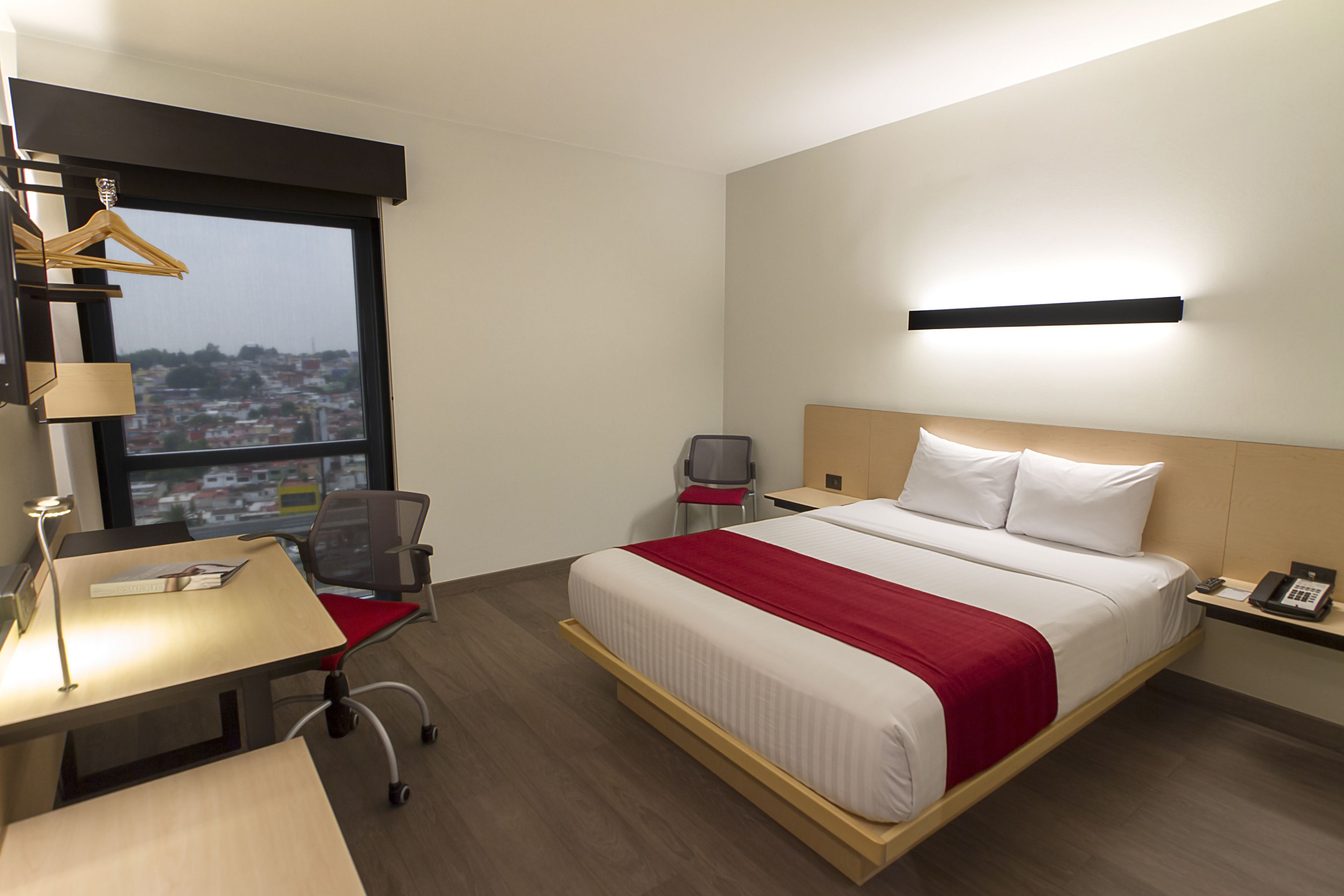 A hotel room with a large bed, a desk with a chair, a small table, a chair, a bedside table with a phone, and a window with a view of a cityscape.