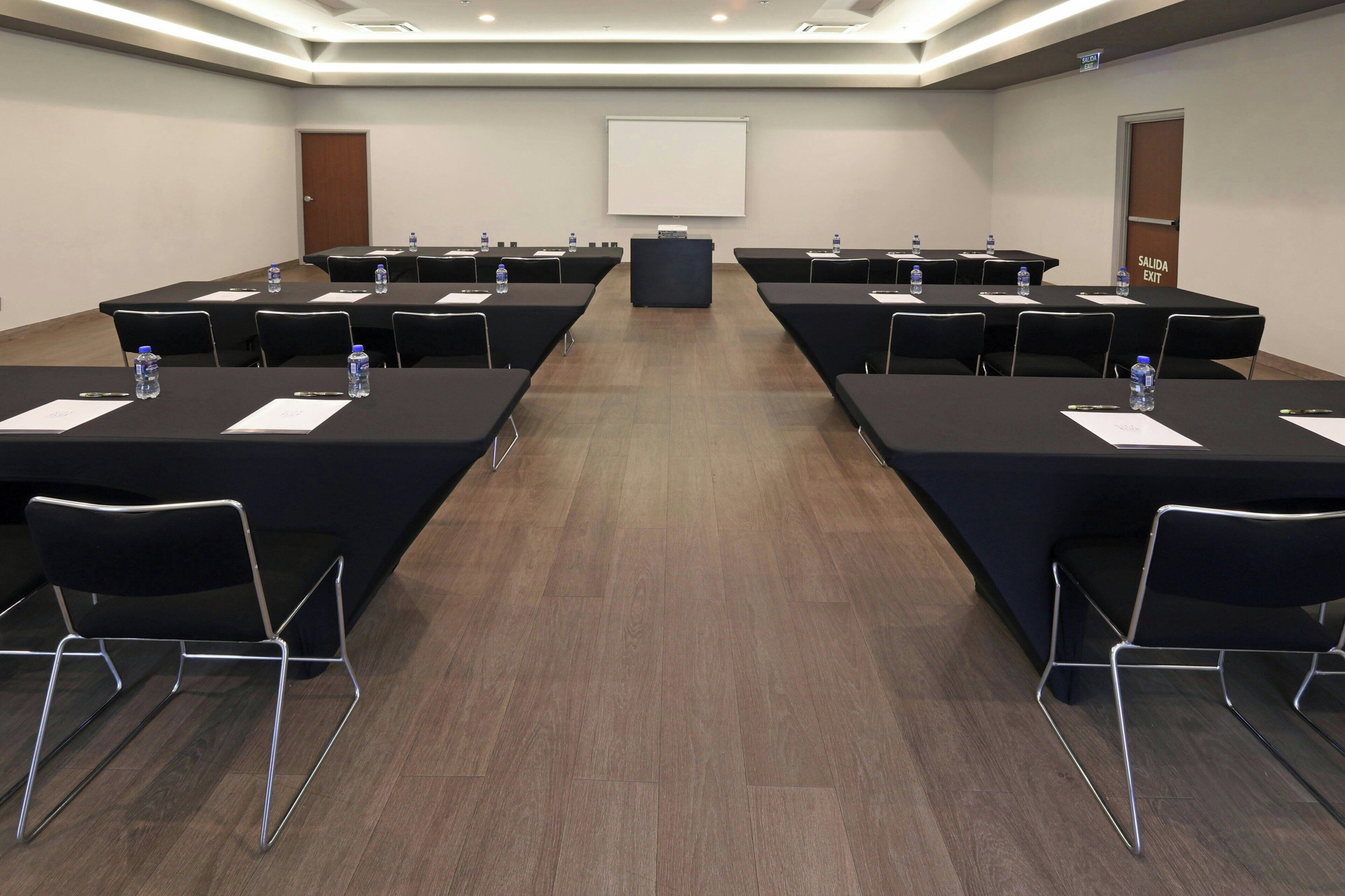 A conference room with long tables, chairs, water bottles, and a projector screen.