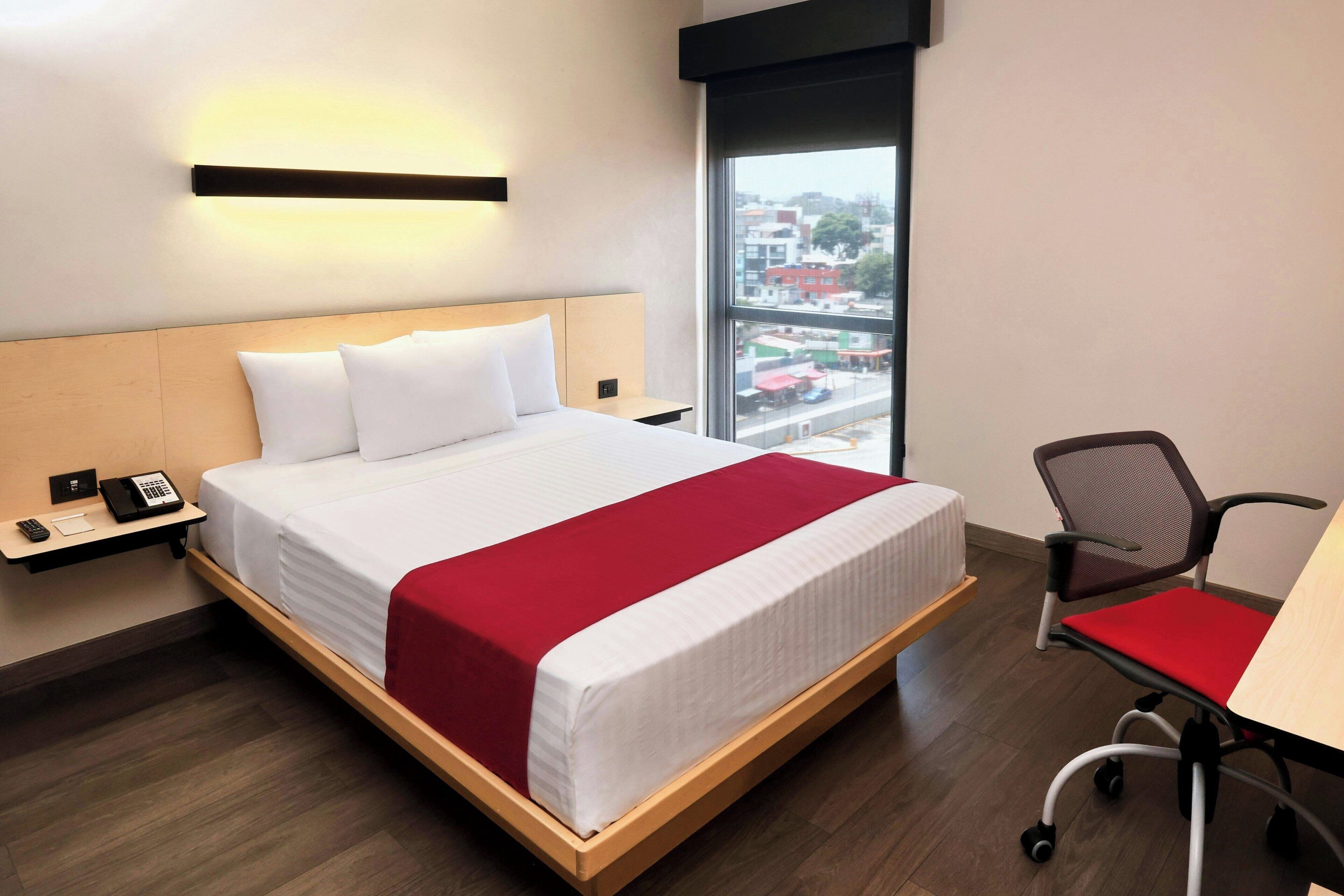 Une chambre d’hôtel équipée d’un lit, d’un bureau, d’une chaise et d’une grande fenêtre donnant sur la ville.
