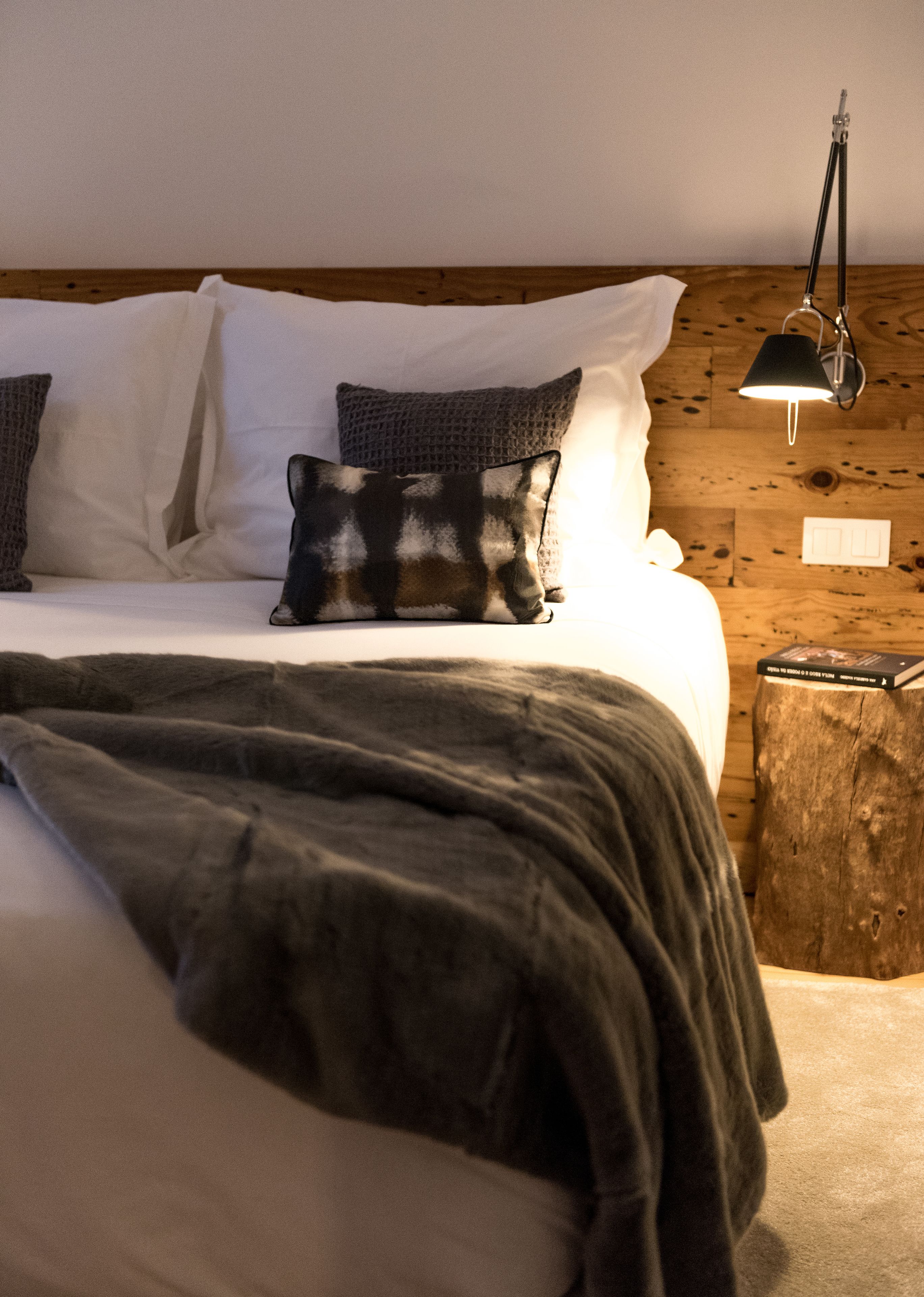 Una cama bien tendida con un edredón oscuro, dos almohadas y una mesita de noche con una lámpara y un libro.