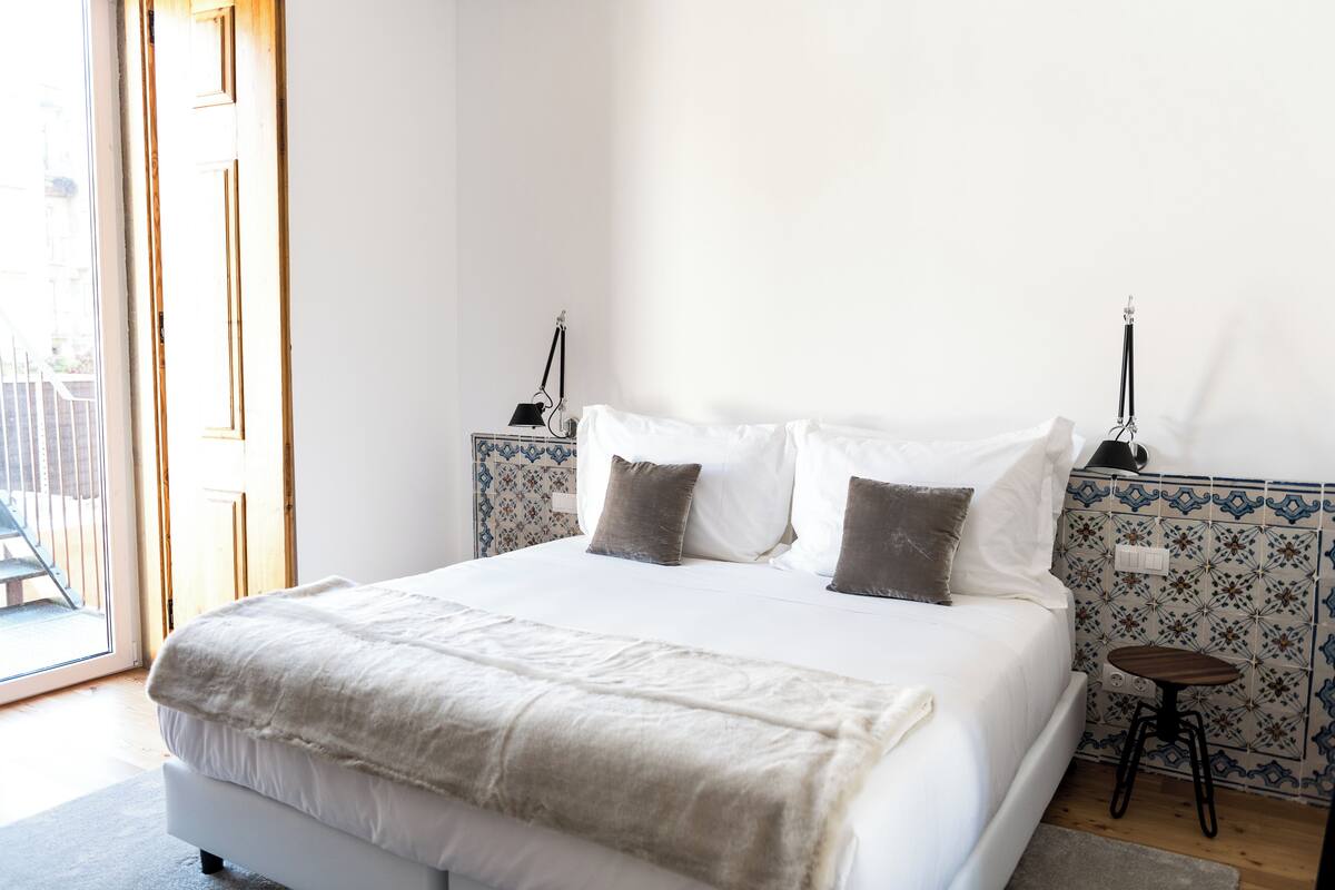 A neatly made bed with a white duvet and two pillows, a bedside table with a lamp, and a patterned tile wall.