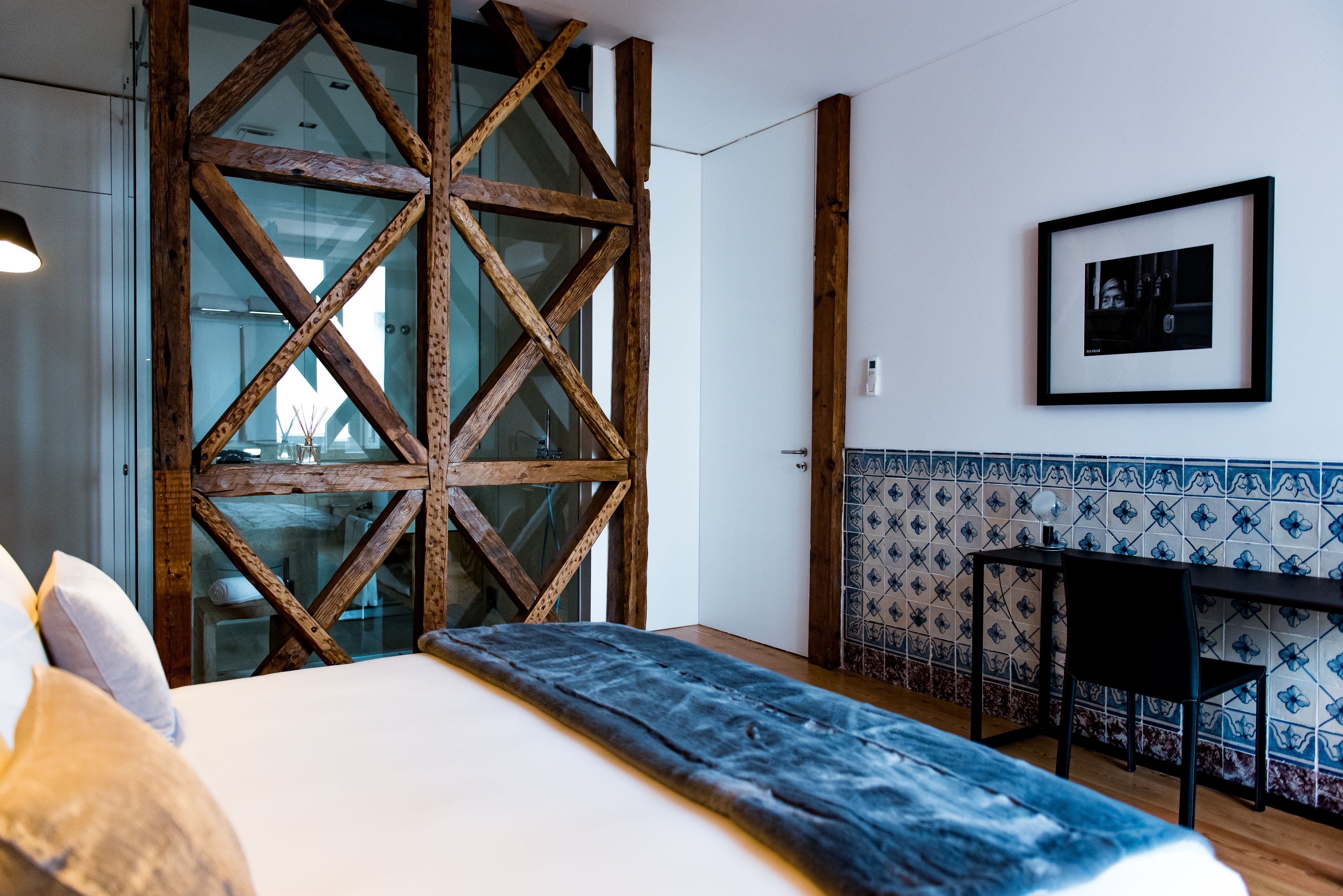 A hotel room with a large bed, a wooden partition, a small table with a chair, and a tiled wall with a framed picture.