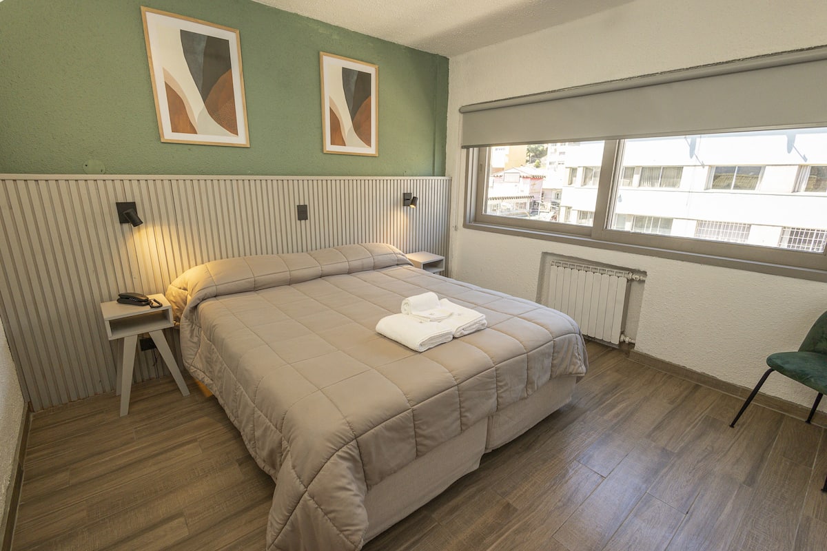 A hotel room with a bed, a chair, a nightstand, and a window with a view of buildings.