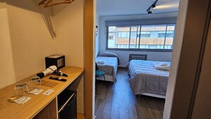 A compact hotel room with a kitchenette, two single beds, a large window, and a hanging clothes rack.