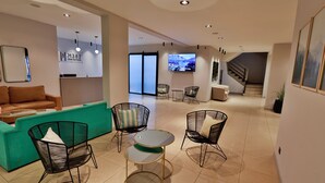 A modern hotel lobby with a teal reception desk, black wicker chairs, a small round table, and a TV mounted on the wall.