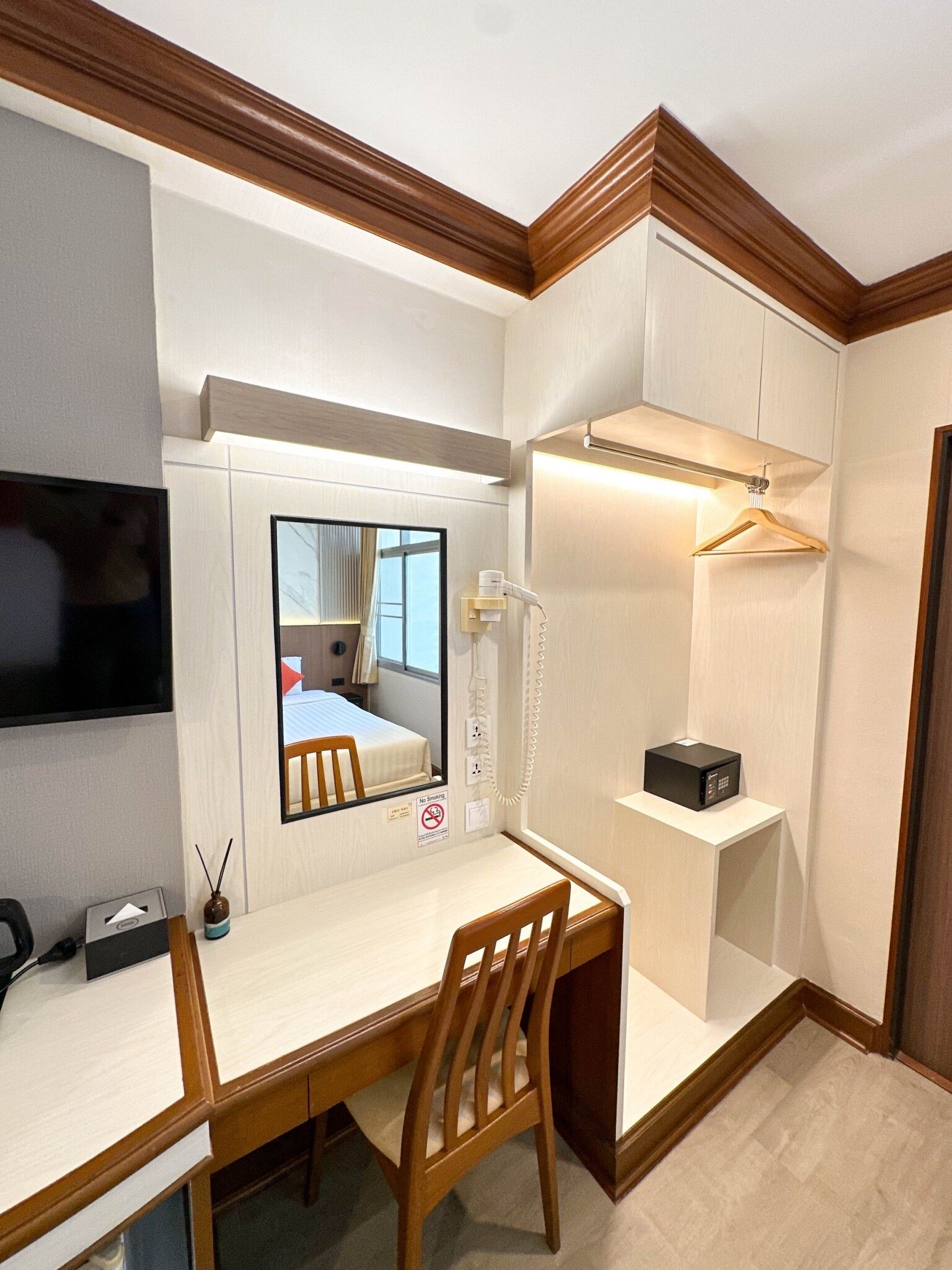A hotel room with a desk, chair, a mirror, a TV, a telephone, a microwave, and a hanging light fixture.