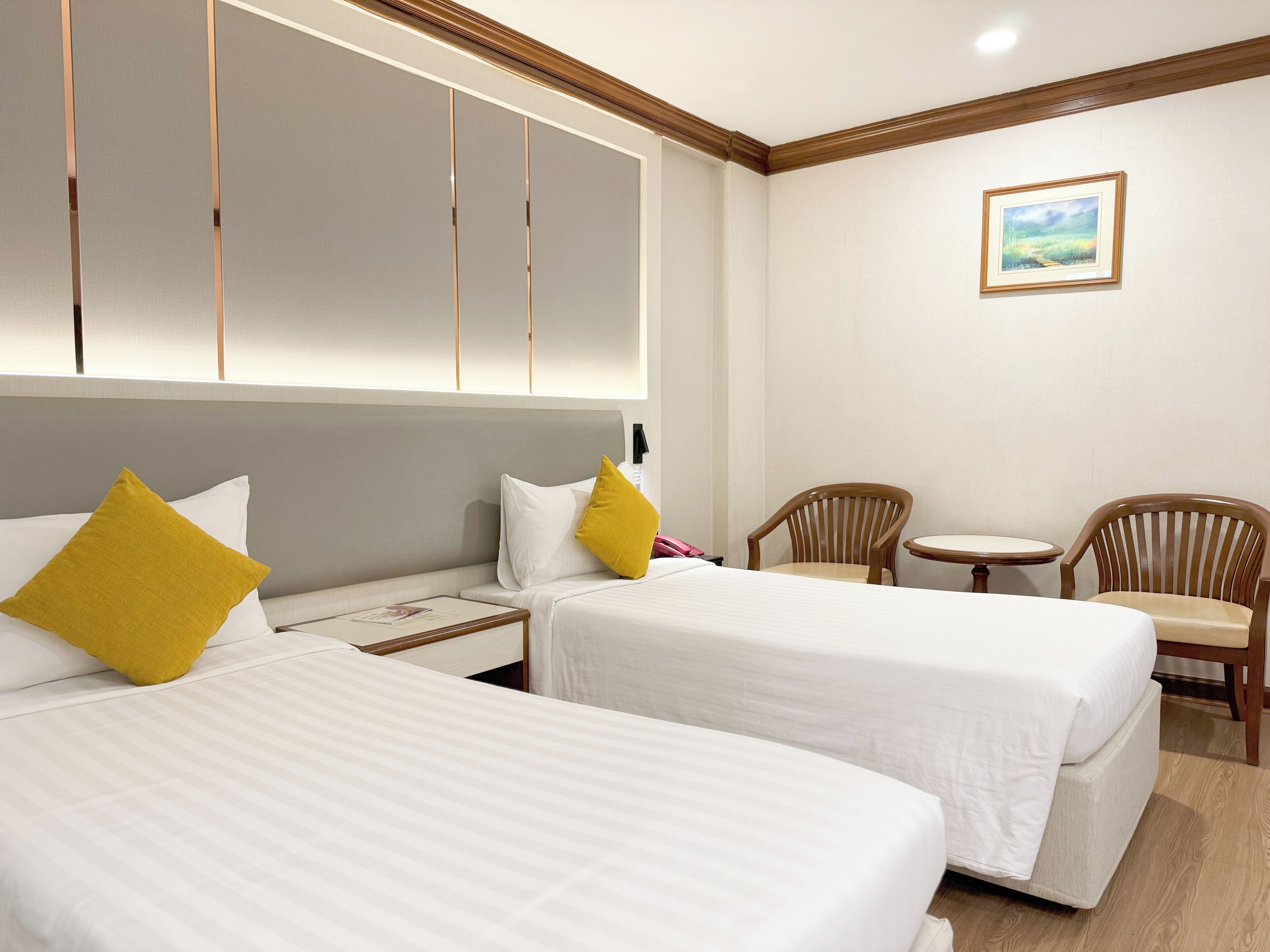 A hotel room with two beds, a small table, two chairs, a painting on the wall, and a wooden ceiling trim.