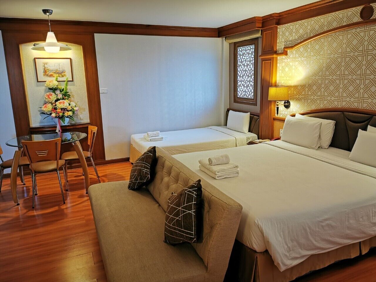 A hotel room with two beds, a dining table with chairs, a small table with a vase of flowers, and a decorative wall panel.