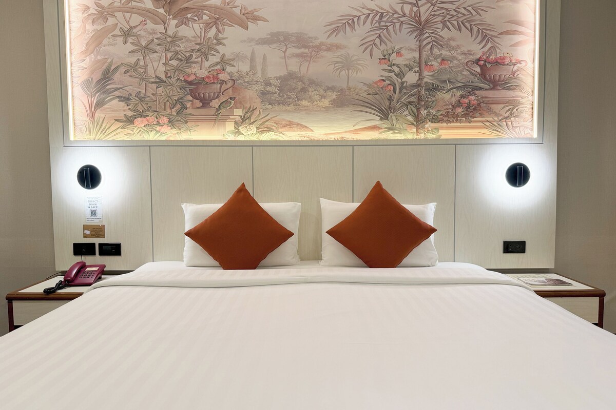 Ein Hotelzimmer mit einem großen Bett, zwei Kissen, einem Nachttisch mit Telefon und einem Wandbild, das eine tropische Szene zeigt.