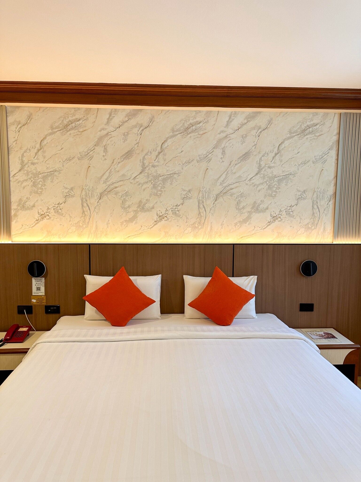 Une chambre d’hôtel avec un grand lit, deux oreillers, une table de chevet, un téléphone et un mur au motif rappelant le marbre.