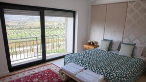 A bedroom with a large window, a bed with patterned bedding, a red and white rug, and a small bedside table with a lamp. - Quinta da Terrincha (Torre de Moncorvo)