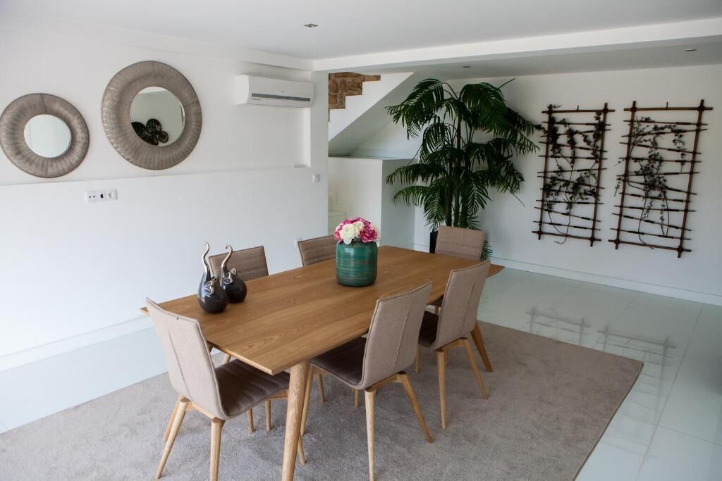 A dining area with a wooden table, beige chairs, and a vase with flowers. There are two wall-mounted mirrors and a climbing plant.