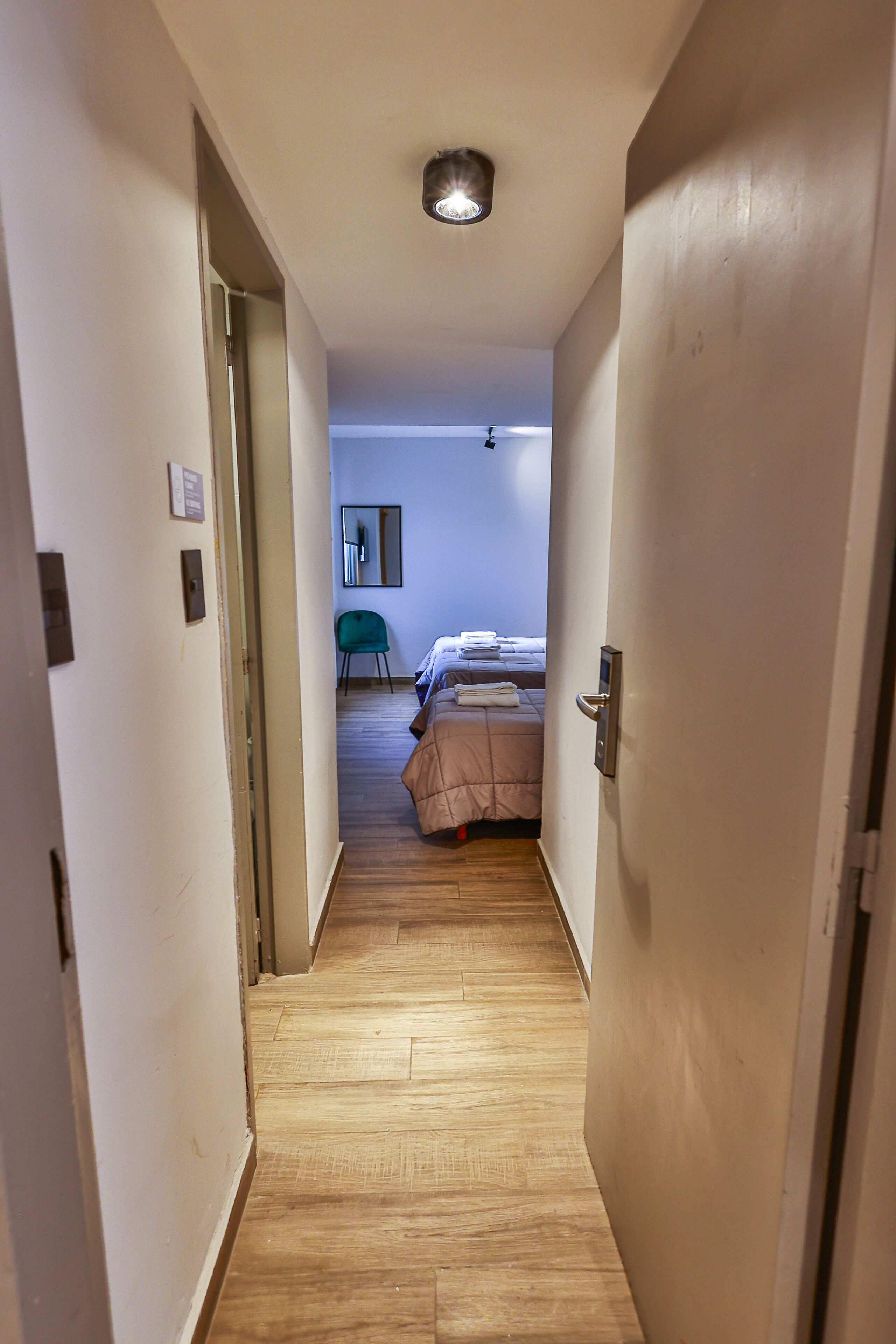 A narrow hallway with wooden flooring leading to a bedroom with a bed and a chair.
