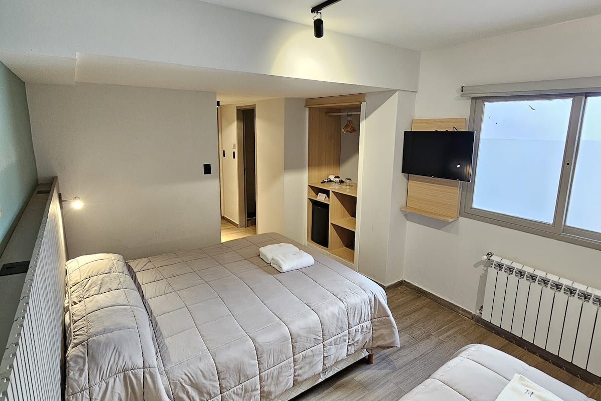 A modern bedroom with a large bed, a smaller sofa, a TV mounted on the wall, and a window providing natural light.