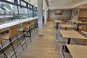 Un'area pranzo moderna con tavoli e sedie alti, ampie finestre e un televisore a schermo piatto appeso al muro.