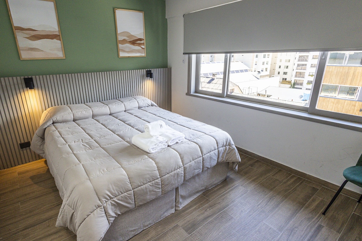 A neatly made bed with a grey quilt, a wooden floor, and a window with a view of buildings outside.