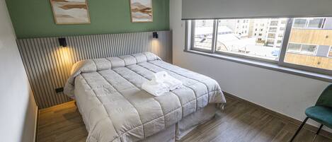 Un lit bien fait, recouvert d’une couette grise, un plancher en bois et une fenêtre donnant sur des bâtiments à l’extérieur.