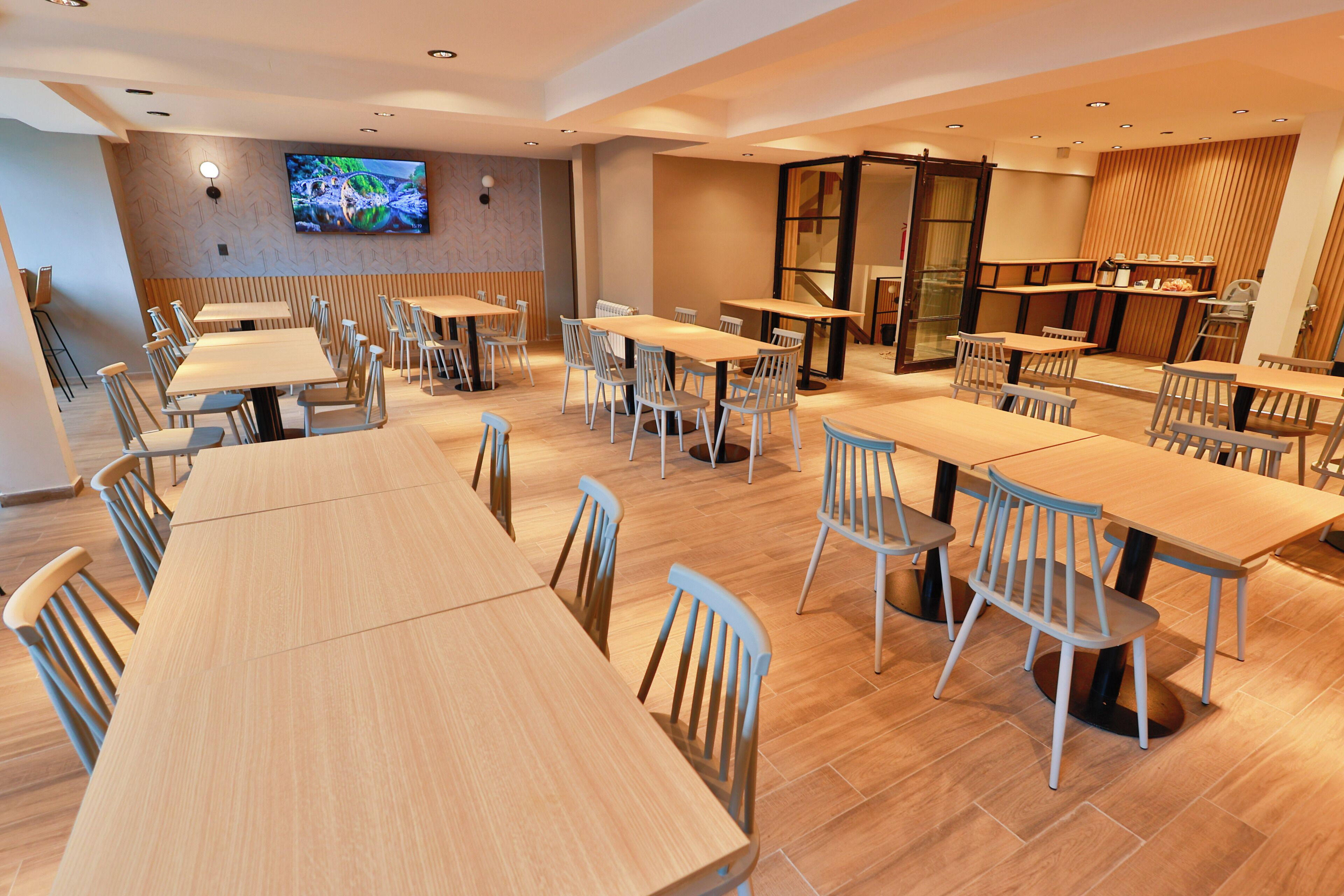 Un grand espace de salle à manger avec plusieurs tables et chaises en bois, un téléviseur fixé au mur et un coin cuisine visible à travers une porte ouverte.