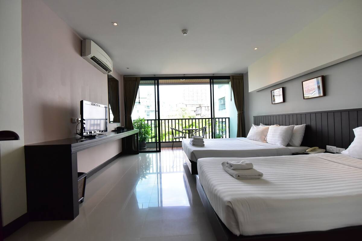 A hotel room with two beds, a desk with a computer, a TV, and a balcony with a view of buildings.