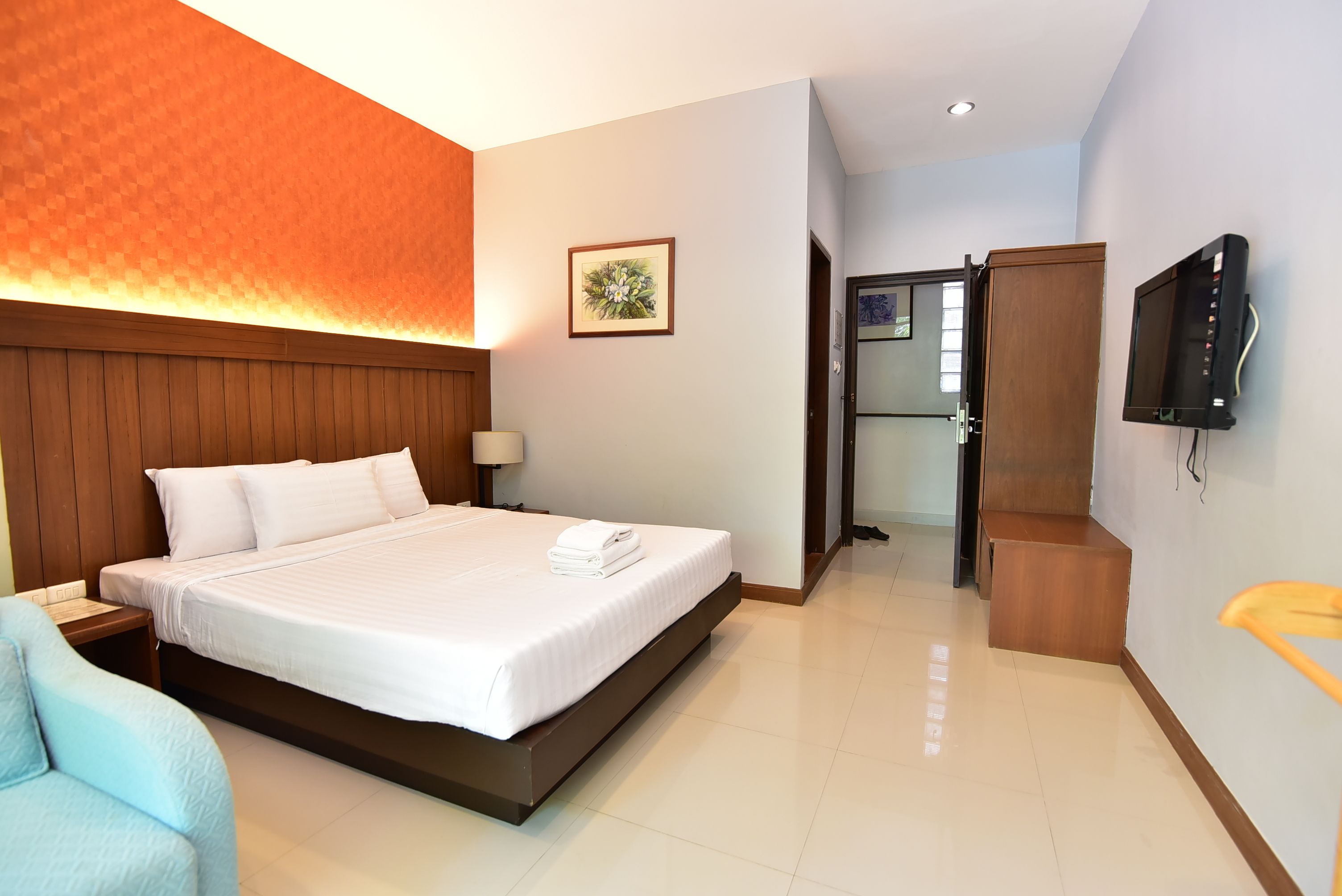 A hotel room with a bed, a TV, a wooden headboard, a nightstand, a chair, and a door leading to another room.
