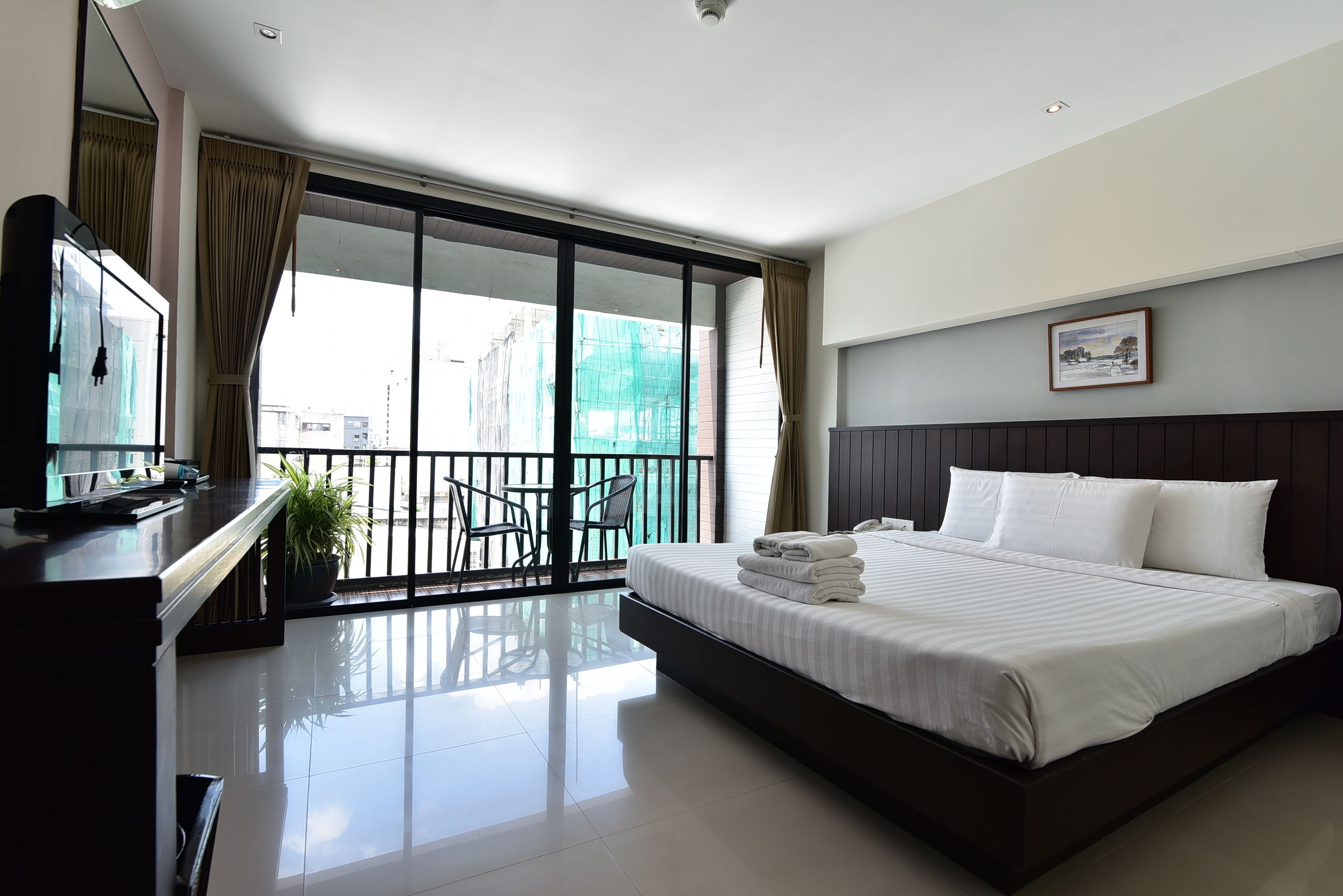 A hotel room with a large bed, a desk with a TV, a balcony with chairs, and a view of buildings outside.