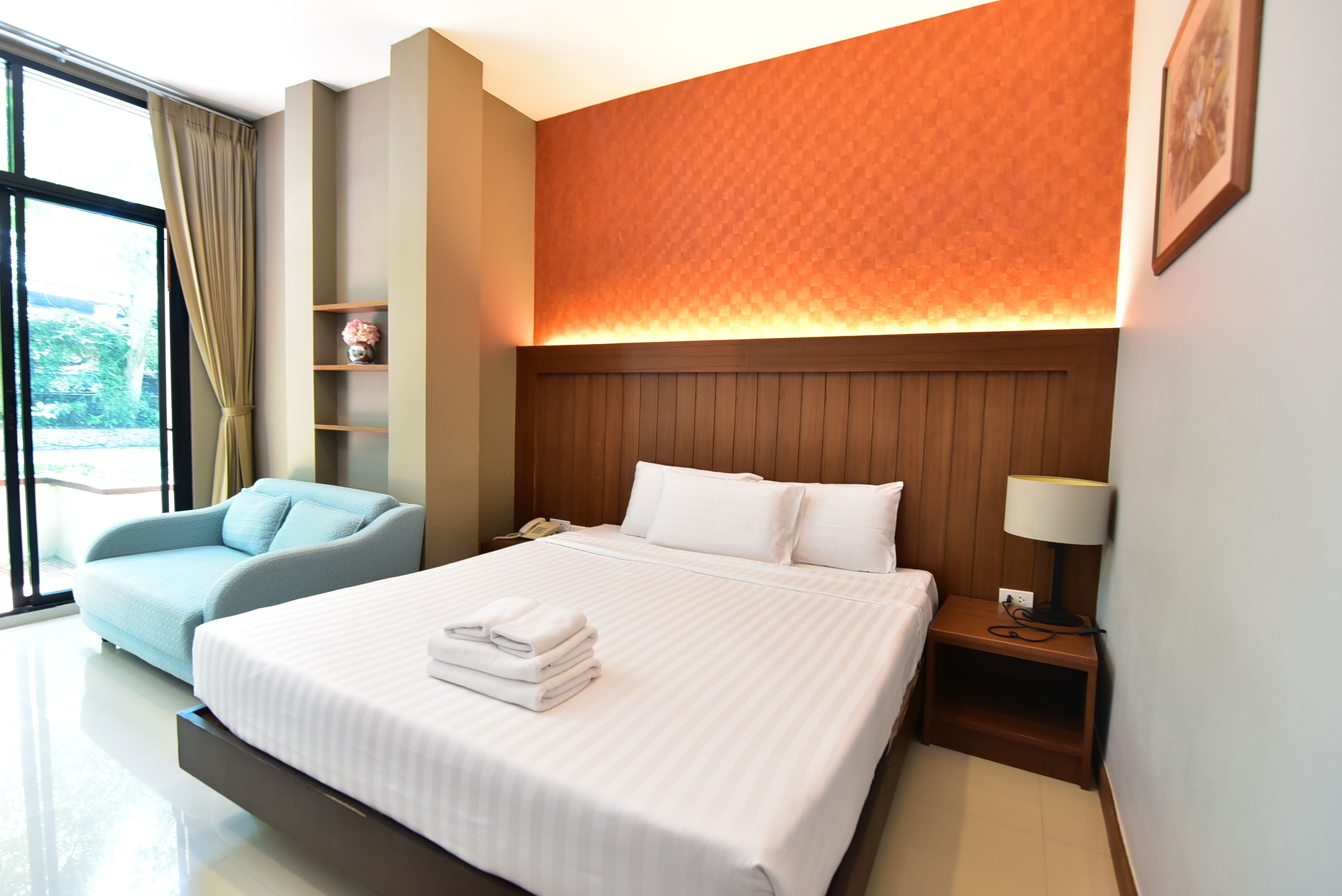 A hotel room with a large bed, a blue armchair, a bedside table with a lamp, and a view of greenery outside.